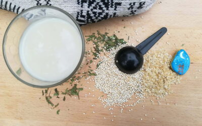 vaso con leche de quinoa sobre mesa de madera junto a copos y granos de quinoa y un gorro con motivos andinos